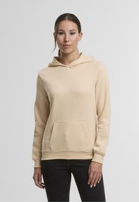 Sudadera beige hecha de un tejido suave y texturizado con bolsillo frontal y capucha con cordón, combinada con jeans negros. Diseño simple y versátil.