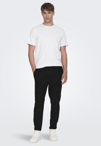 Camiseta blanca de algodón y pantalones de chándal negros ajustados, con una banda elástica en la cintura. El modelo lleva zapatillas blancas, con un fondo neutro.