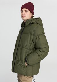 Oliwkowa kurtka puchowa z kapturem, czarnym zamkiem błyskawicznym i kieszeniami bocznymi. Posiada pikowaną strukturę oraz regulowany sznurek na dole.