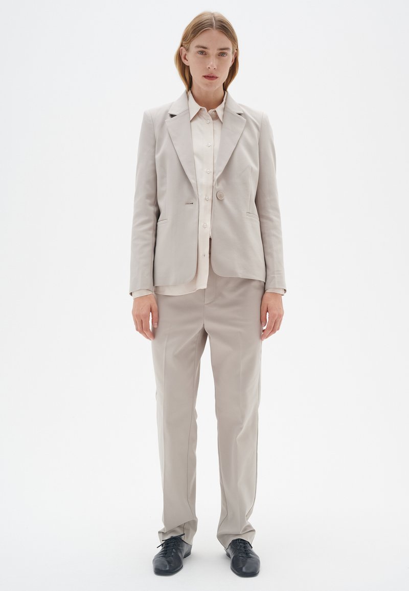 Helles graues, maßgeschneidertes Blazer-Jackett und passende Hose, kombiniert mit einem hellen Hemd mit Knöpfen und abgerundet mit schwarzen Schnürschuhen.