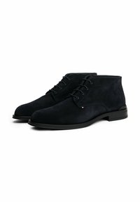 Un paio di stivali alla caviglia da uomo in suede nero con lacci sul davanti e tacco basso, mostrati fianco a fianco su sfondo bianco.