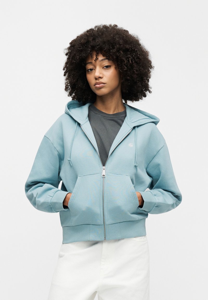 Femme aux cheveux bouclés portant un sweat à capuche bleu clair zippé sur un t-shirt sombre et un pantalon blanc, debout les mains dans les poches devant un fond blanc.