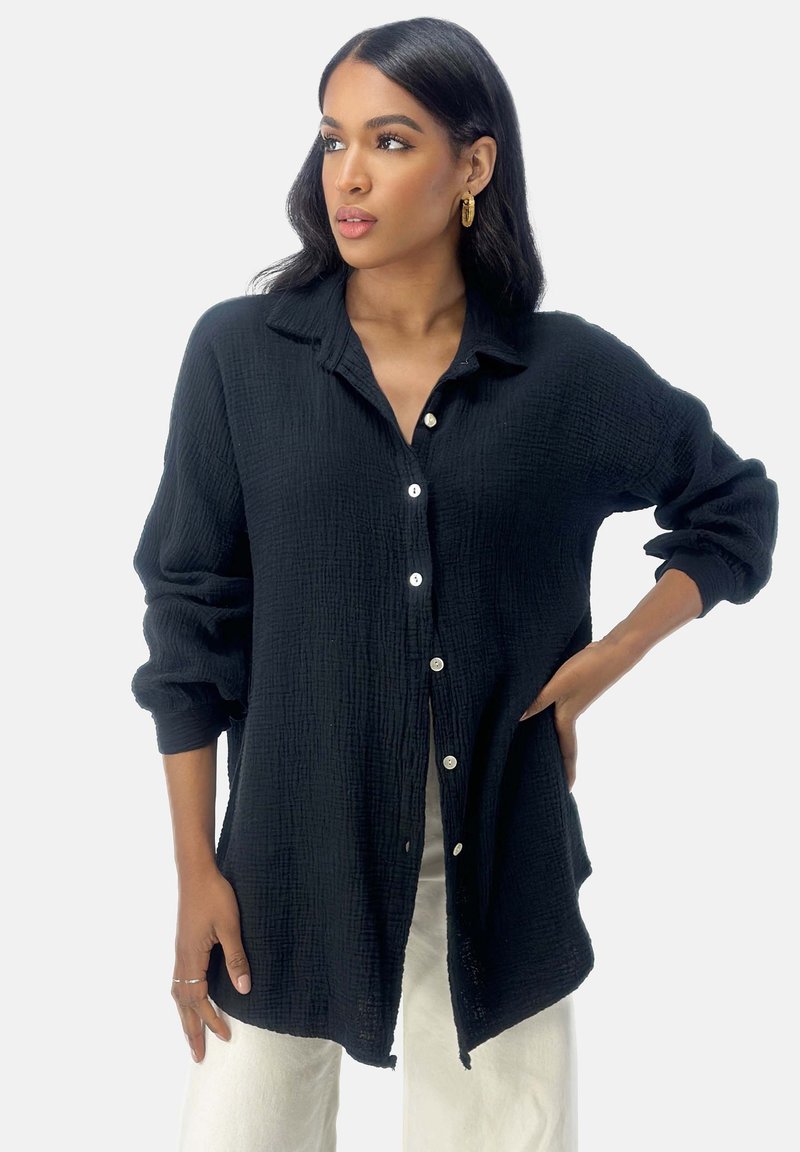 Camisa negra de botones fabricada en una tela texturada, con un diseño de cuello y mangas enrolladas. Notables botones blancos a lo largo del frente.