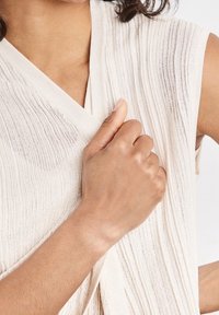 Maglietta a costine beige senza maniche, con una leggera lucentezza e tessuto testurizzato. Una mano tiene delicatamente il tessuto all'altezza del collo.