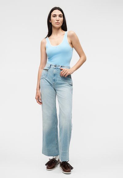 Γυναίκα όρθια, φορώντας ένα γαλάζιο αμάνικο τοπ, τζιν με φαρδιά μπατζάκια και ψηλή μέση με ανοιχτό χρώμα πλύσης, και καφέ μοκασίνια με φούντες.