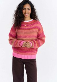 Kobieta o długich, kręconych włosach, uśmiechająca się, ubrana w różowy i pomarańczowy sweter w paski oraz ciemne spodnie, stojąca na tle białego tła.