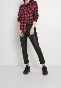 Red and black checkered flannel shirt with buttoned front, two chest pockets; paired with black leather tapered pants and black sneakers.