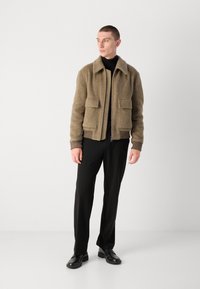 Braune, strukturierte Jacke mit Kragen, zwei Vordertaschen, gerippten Bündchen und einem Reißverschluss, kombiniert mit schwarzen Hosen und Schuhen.