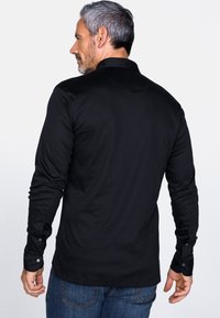 Schwarzes Langarmshirt aus glattem Stoff, ausgestattet mit einem Kragen, knöpfbaren Manschetten und einem eleganten, figurbetonten Schnitt, der auf der Rückseite sichtbar ist.