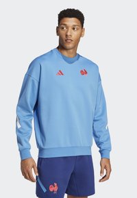 Vaaleansininen collegepaita, joka on valmistettu pehmeästä kankaasta, jossa on punainen kukko-logo ja valkoiset yksityiskohdat hihoissa, yhdistettynä tummansinisiin shortsihin.