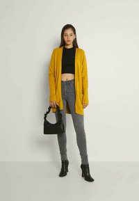 Vrouw met een mosterdgele cardigan, zwart cropped topje, grijze spijkerbroek, zwarte enkellaarsjes, en een zwarte handtas met een gedraaide handgreep.