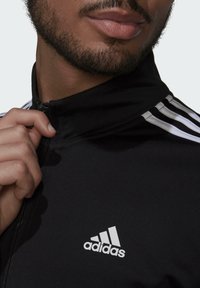 Chaqueta atlética negra con cremallera y acentos en franjas blancas en los hombros, con un logo blanco de Adidas en el lado izquierdo del pecho, y un cuello alto.