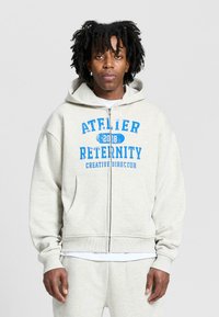 Jasnoszara, zapinana na zamek bluza z miękkiego materiału. Posiada niebieski nadrukowany tekst: "ATELIER RETERNITY 2018 DYREKTOR KREATYWNY." W zestawie kieszenie kangurze.