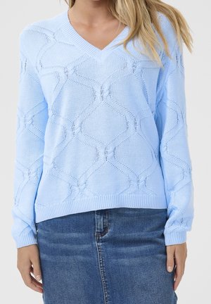 Femme portant un pull en maille bleu clair à col en V avec des motifs géométriques et des manches longues, associé à une jupe en jean bleue.