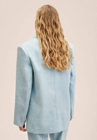 Veste oversize bleu clair en tissu texturé, avec poignets à trois boutons et fente centrale au dos. Portée sur des cheveux ondulés.