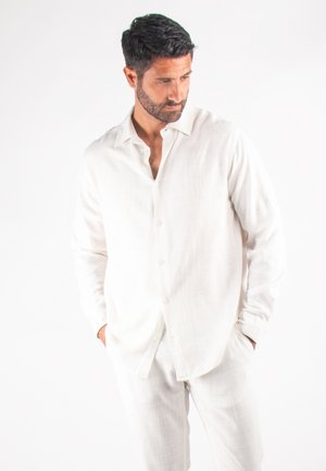 Chemise blanche en lin à boutons avec manches longues, coupe décontractée et surface texturée, associée à un pantalon en lin assorti, se tenant devant un fond uni.