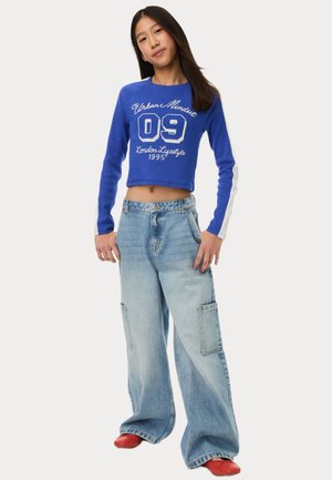Blauwe cropped sweatshirt met witte tekst, gecombineerd met lichtblauwe hoge taille jeans met zijzakken. Rode schoenen maken de outfit af.