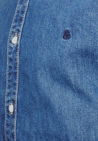 Detailní záběr na modrou denimovou košili s bílými knoflíky a tmavě modrým vyšitým ampersandem v blízkosti hrudní části.