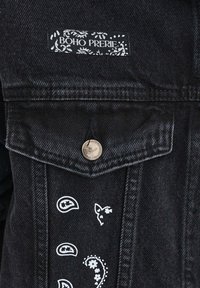 Černá džínová bunda s označením "BOHO PRERIE", kovovým knoflíkem a bílými paisley a květinovými potisky v oblasti kapsy.