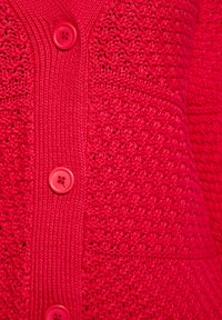 Cardigan rosso lavorato a maglia con un design testurizzato, caratterizzato da un pannello centrale liscio e grandi bottoni coordinati. Ingrandimento del dettaglio del tessuto.