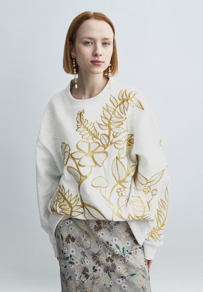 Donna con capelli rossi corti che indossa lunghi orecchini di perle, una felpa bianca con ricami floreali dorati e una gonna a motivo floreale.