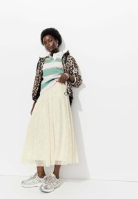 Veste à imprimé léopard sur un polo à rayures vertes et roses, jupe en dentelle blanche et baskets argentées avec des lacets texturés. Silhouette lisse, look superposé.