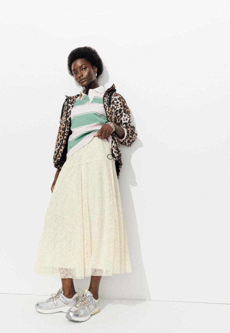 Veste à imprimé léopard sur un polo à rayures vertes et roses, jupe en dentelle blanche et baskets argentées avec des lacets texturés. Silhouette lisse, look superposé.