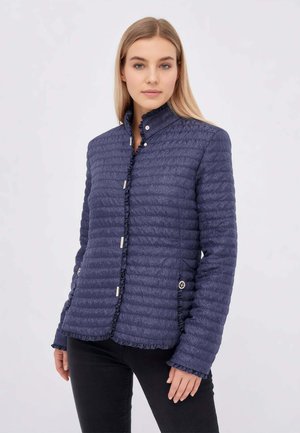 Donna che indossa una giacca trapuntata blu navy con bottoni e bordi arricciati e pantaloni neri, in piedi davanti a uno sfondo chiaro e uniforme.