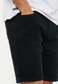 Homme portant un short noir retroussé avec une main dans la poche, associé à une chemise blanche affichant une étiquette "THREADBARE".