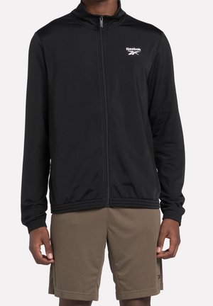 Homme portant une veste zippée noire Reebok et un short marron, debout avec les bras détendus le long du corps devant un fond uni.