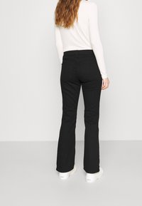 Jean noir évasé à taille haute, en denim résistant, avec deux poches arrière. Associé à des baskets blanches.