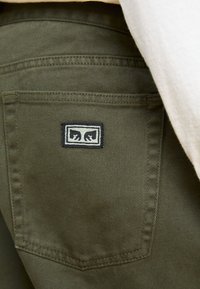 Pantalons en denim vert olive avec une poche arrière montrant une étiquette rectangulaire en logo noir et blanc, cousue sur le tissu.