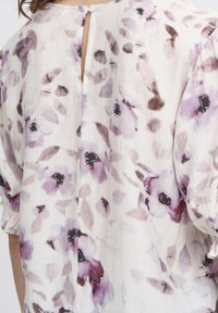Blusa floreale con sfondo bianco e segni viola e scuri. Apertura a occhio di chiave sul retro con chiusura a bottone. Tessuto leggero e strutturato.