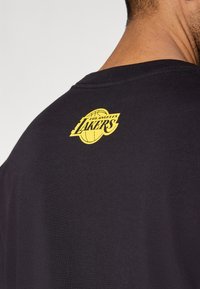 Μαύρο βαμβακερό T-shirt με κίτρινο λογότυπο "Los Angeles Lakers" στην πλάτη κοντά στο λαιμόκοψη. Μαλακή υφή, σχέδιο με στρογγυλή λαιμόκοψη.