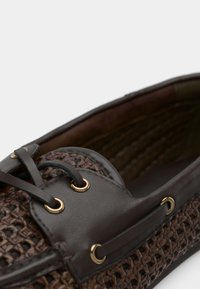 Chaussure tissée marron avec des accents en cuir, dotée d'œillets et de lacets dorés, et d'une surface texturée pour plus d'adhérence. L'intérieur est doublé d'un matériau foncé.