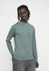 Ανοιχτοπράσινο φούτερ από ύφασμα με μείγμα βαμβακιού, με λαιμόκοψη crew, μακριά μανίκια και ριμπ τελείωμα. Συνδυάζεται με γκρι τζιν.