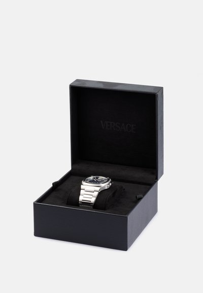 Reloj Versace en una caja negra con interior de terciopelo; presenta una correa plateada, esfera azul y caja de acero inoxidable. Logo grabado en el interior de la caja.