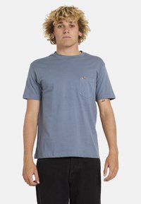 Camiseta de algodón azul con manga corta, cuello redondo y un bolsillo en el pecho que presenta una pequeña etiqueta roja con el logo. Diseño simple y clásico.