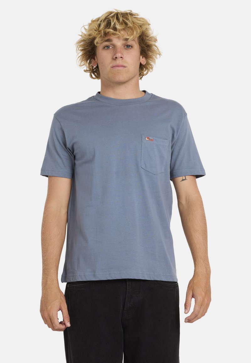 Camiseta de algodón azul con manga corta, cuello redondo y un bolsillo en el pecho que presenta una pequeña etiqueta roja con el logo. Diseño simple y clásico.