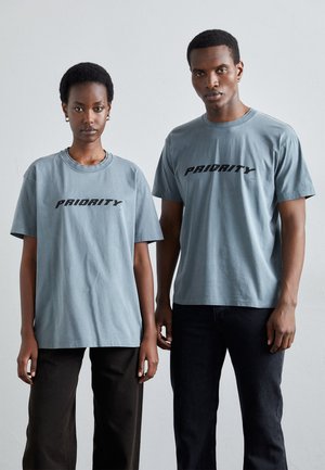 Gray cotton t-shirts featuring "PRIORITY" graphic in black print. Relaxed fit with short sleeves, paired with dark trousers.