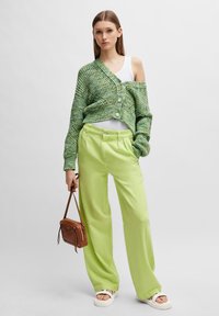 Grønn strikket cardigan, hvit tanktopp, limegrønne vide bukser og en brun crossover-veske. Har knapper og en avslappet passform, med flate sandaler.