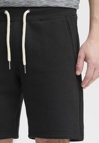Shorts en coton noir avec une taille côtelée et un cordon de serrage crème. Ils comportent des poches latérales et une texture lisse avec des accents de couture.