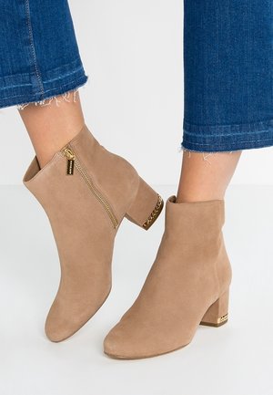 Bottines en daim couleur camel avec fermeture éclair dorée et détail chaîne sur talons blocs, portées avec un jean bleu à ourlet effiloché.
