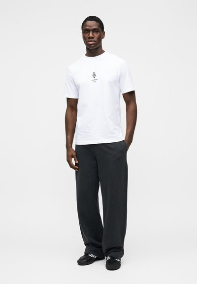 Άσπρο μπλουζάκι με ένα μικρό μαύρο γραφικό στο κέντρο, συνδυασμένο με άνετο μαύρο παντελόνι και μαύρα παπούτσια με λευκές λεπτομέρειες.