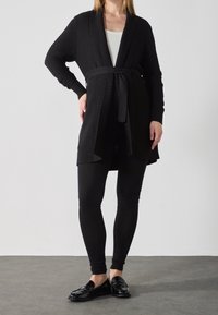 Svart stickad cardigan med bälte, ribbad textur, långa ärmar, öppen front, matchad med svarta leggings och loafers på ett grått golv.