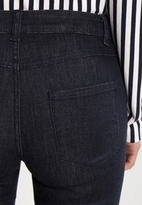 Pantalones de mezclilla azul oscuro con una cintura alta, con un solo bolsillo trasero. La tela tiene una textura suave y detalles de costura sutiles.