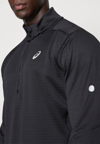 Svart atletisk pullover med halvdragkedja, strukturerat tyg, reflexlogotyp på bröstet och en liten reflexpunkt på ärmen.