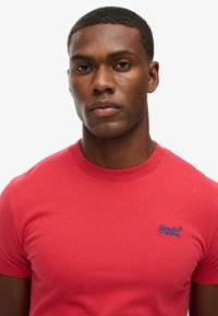 Camiseta corta roja de algodón con cuello redondo. Presenta un pequeño logotipo bordado azul en el lado superior derecho.