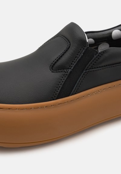 Melna ādas slip-on kurpe ar elastīgām sānu panelēm un brūnu gumijas zoli. Ir gluda virsma ar šuvju detaļām. Redzams punktiņu oderējums.