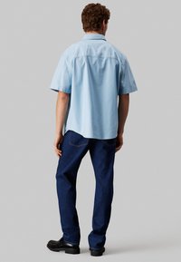 Camicia a maniche corte azzurro chiaro, vista posteriore, abbinata a jeans blu scuro e scarpe nere. La camicia ha una vestibilità comoda e un tessuto leggermente texturizzato.